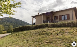 A two-story house with a balcony, surrounded by a grassy hill and a mountain in the background.
