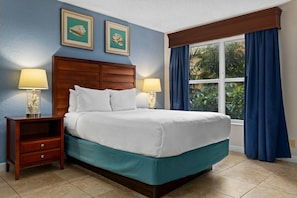 Ein Hotelzimmer mit einem Bett, zwei Nachttischlampen, einem Nachttisch, einem Fenster mit Vorhängen und zwei gerahmten Bildern an der Wand.