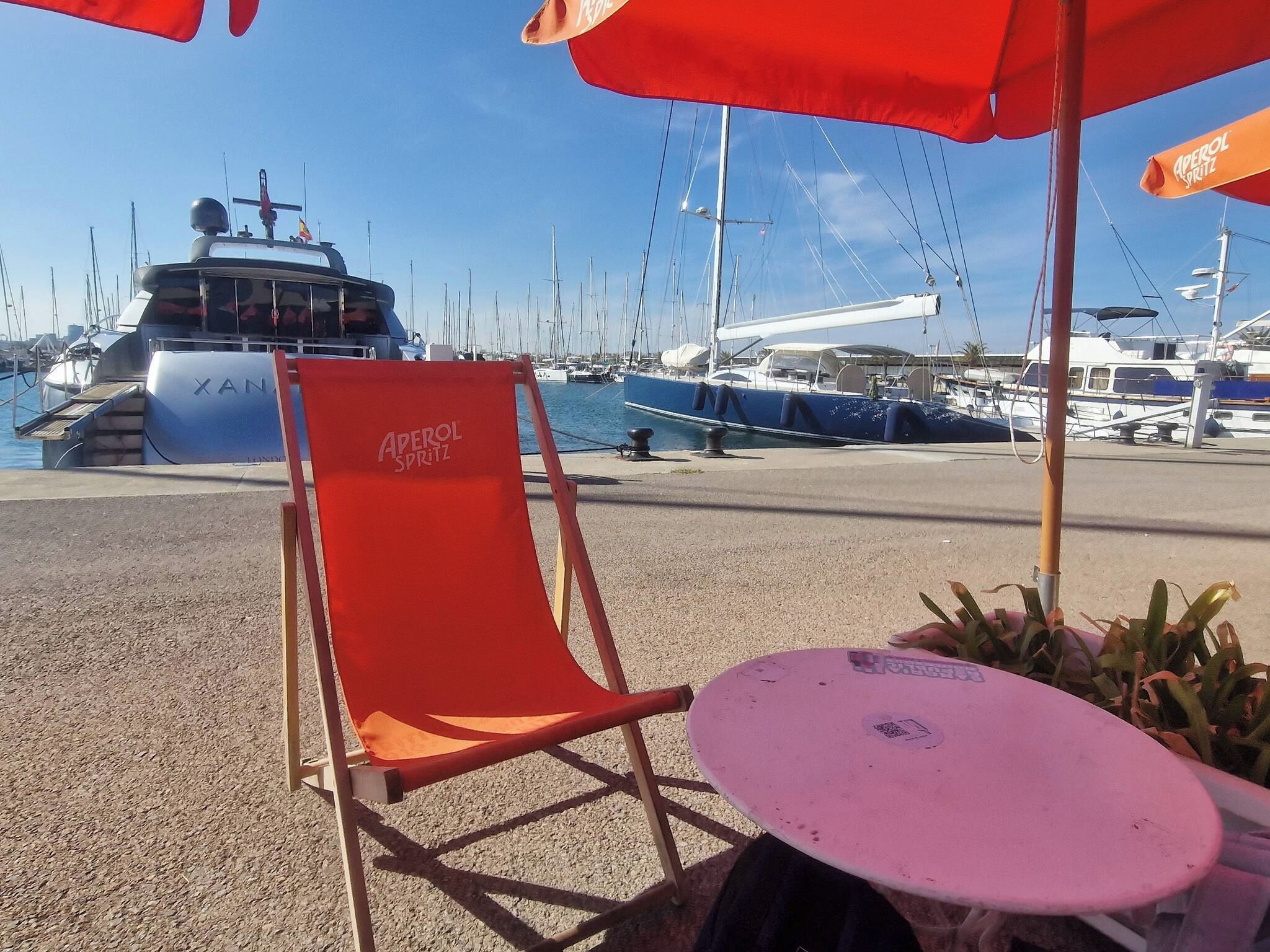 Un port esportiu amb vaixells, una cadira vermella amb el text 'APEROL SPRITZ' i una taula amb superfície rosa.