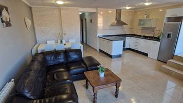 A modern living room with a black leather sofa, a wooden coffee table, and a kitchen in the background.
