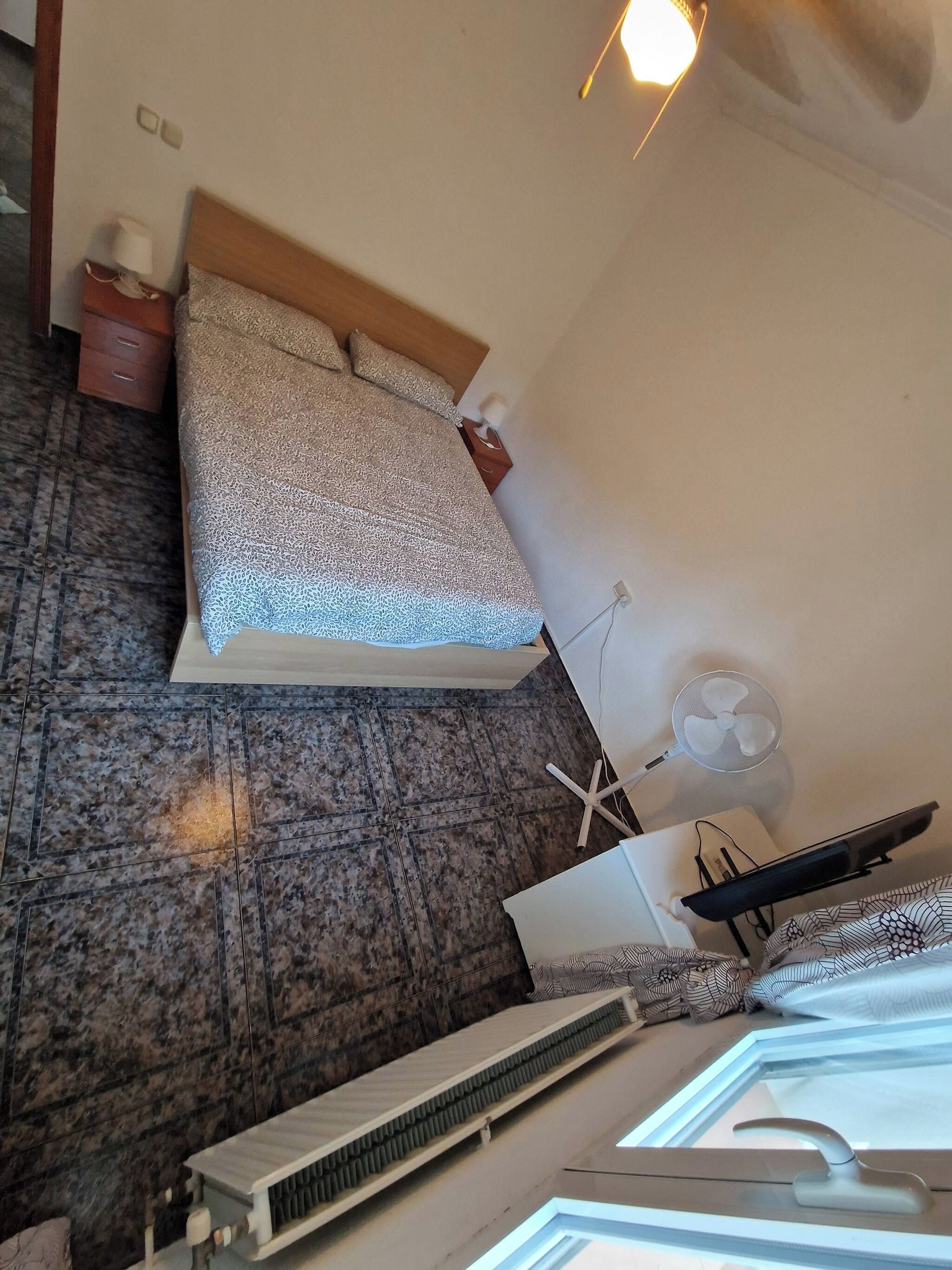 A bedroom with a bed, a nightstand, a lamp, a fan, and a window with blinds.