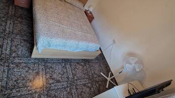 A bedroom with a bed, a nightstand, a lamp, a fan, and a window with blinds.