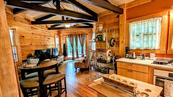A cozy cabin interior with a wooden dining area, kitchen, and a large window with curtains.