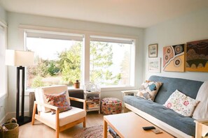 A living room with a sofa, armchair, and a small table. There are windows with a view of greenery outside. - Pool Kayaks BBQ Tides & Tranquility Suite A (Nanoose Bay)
