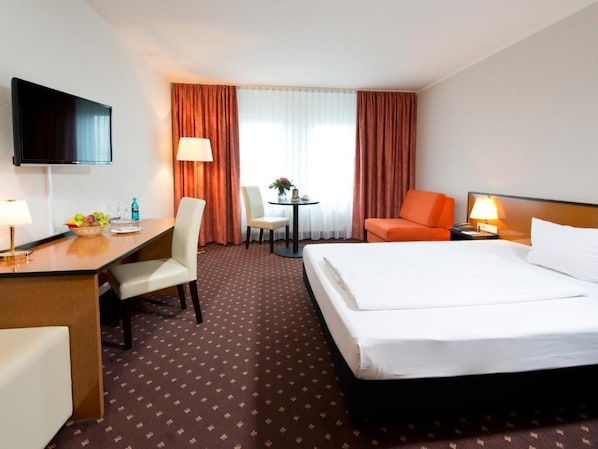Habitación de hotel con cama, escritorio, silla, televisor y una mesita de noche con lámpara y flores.