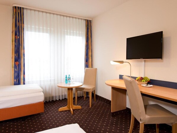 Habitación de hotel con cama, escritorio, silla, televisor, lámpara y una mesita con botellas y fruta.