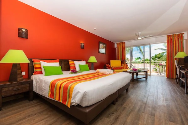 Een hotelkamer met een groot bed, twee nachtkastjes, een bureau en een balkon met uitzicht op palmbomen.