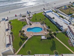 An aerial view of a resort with a swimming pool surrounded by a well-maintained lawn, a beach area with umbrellas, and a parking lot. - Chateau by the Sea 30D | Updated Ocean View Condo | Heated Pool | Direct Beach Access (New Smyrna Beach)