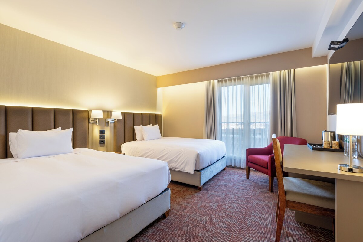 Une chambre d’hôtel avec deux lits, un bureau, une chaise, une fenêtre avec des rideaux et une tête de lit dotée d’un éclairage intégré.
