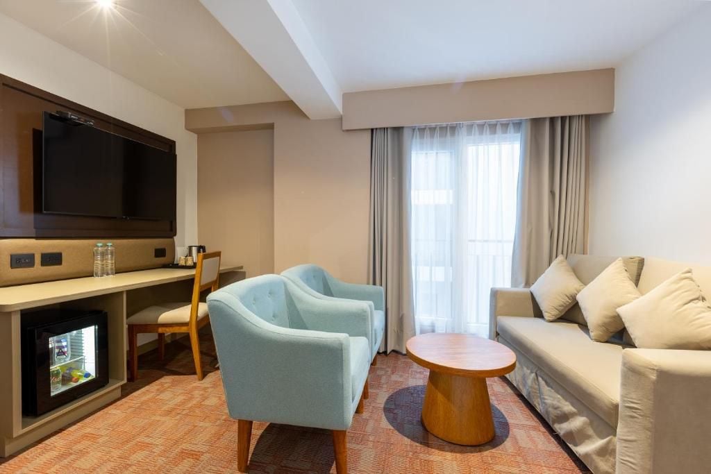 A hotel room with a flat-screen TV, a sofa, two armchairs, a small wooden coffee table, and a carpeted floor.
