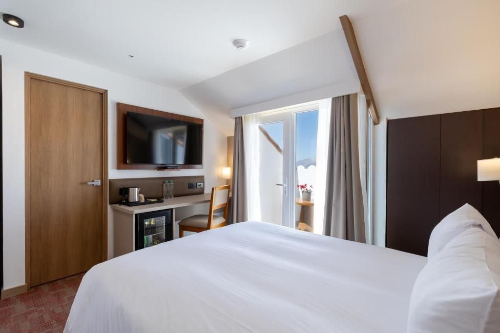 A hotel room with a bed, a desk with a television, a minibar, and a view of the outdoors.
