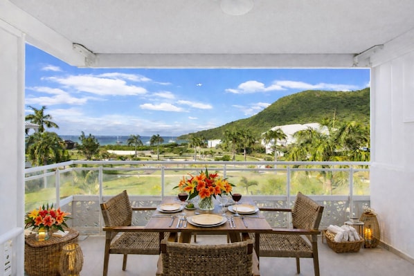 Een balkon met een eettafel, uitkijkend op een kustlandschap met palmbomen en een berg.