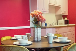A dining area with a round table set for four, white cups and bowls, a white vase with flowers, and a microwave in the background. - Lansing Layover #11 Retro Pink | 1 King (Lansing)