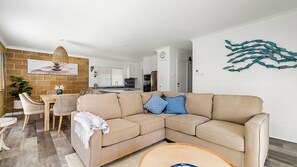 A living room with a beige sectional sofa, a dining area with wicker chairs, and a kitchen in the background. - Easy walker Luxe Villa Walking Distance to Town (Crescent Head)