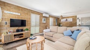 A living room with a brick wall, a flat-screen TV, a beige sofa, and a small wooden coffee table. - Easy walker Luxe Villa Walking Distance to Town (Crescent Head)