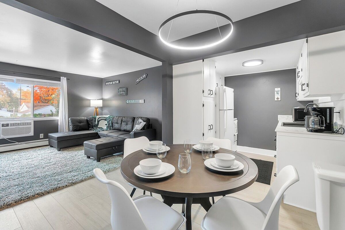 A modern kitchen with white cabinets, a dining area with a round table set for four, and a living area with a sofa and a coffee machine.