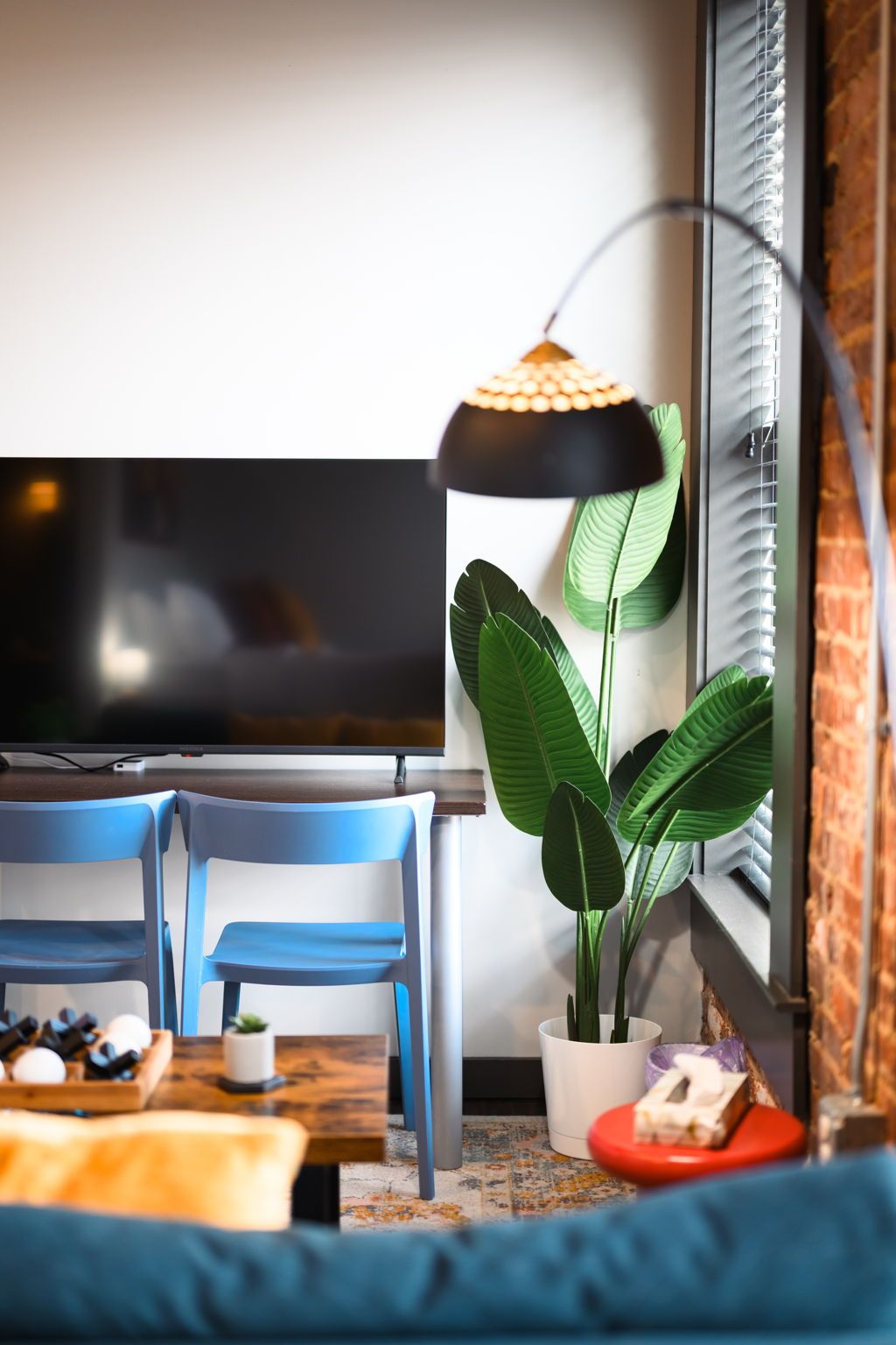 Ein Wohnzimmer mit einem Flachbildfernseher, zwei blauen Sesseln, einem Couchtisch, einer Topfpflanze, einer Stehlampe und einer Ziegelwand.