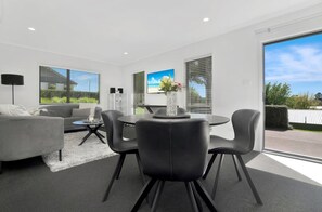 A modern living room with a grey sofa, a round dining table with chairs, a coffee table, and a large window with blinds.