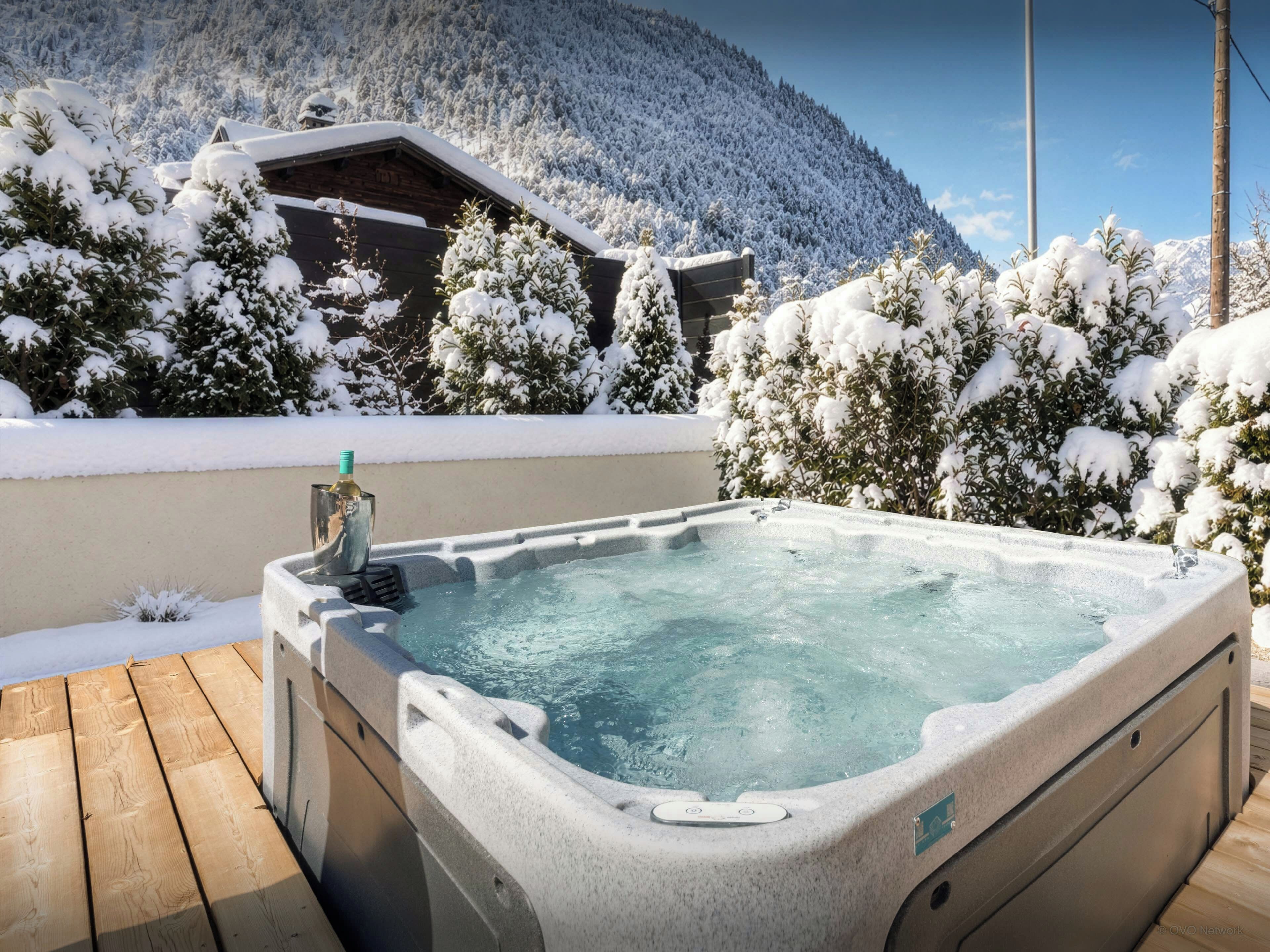 Un bain à remous avec une terrasse en bois, entouré d’arbres couverts de neige et avec une montagne en arrière-plan.