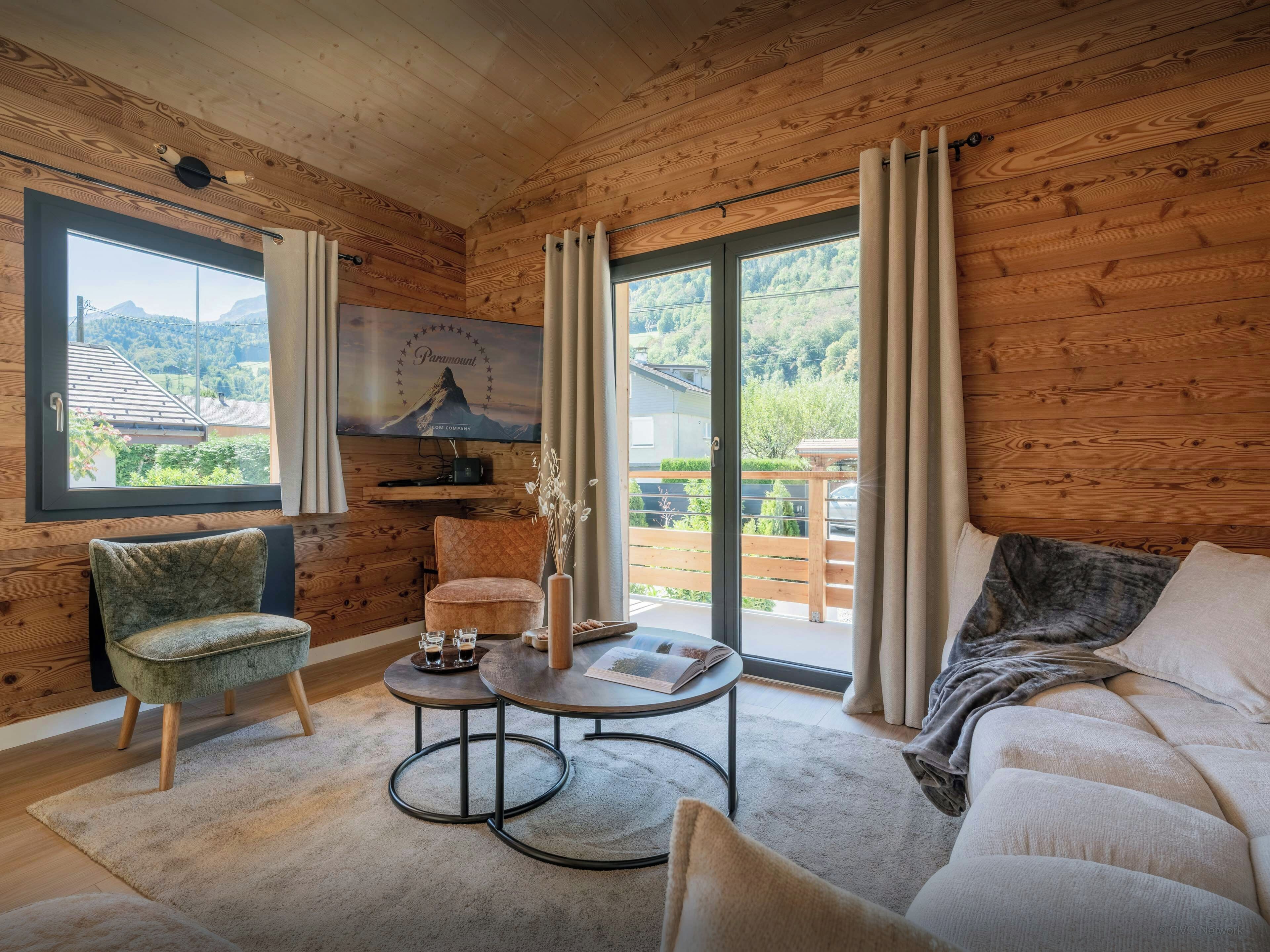 Une pièce chaleureuse avec un décor intérieur en bois, un canapé, un fauteuil, une petite table avec un livre et des lunettes, et un téléviseur fixé au mur.