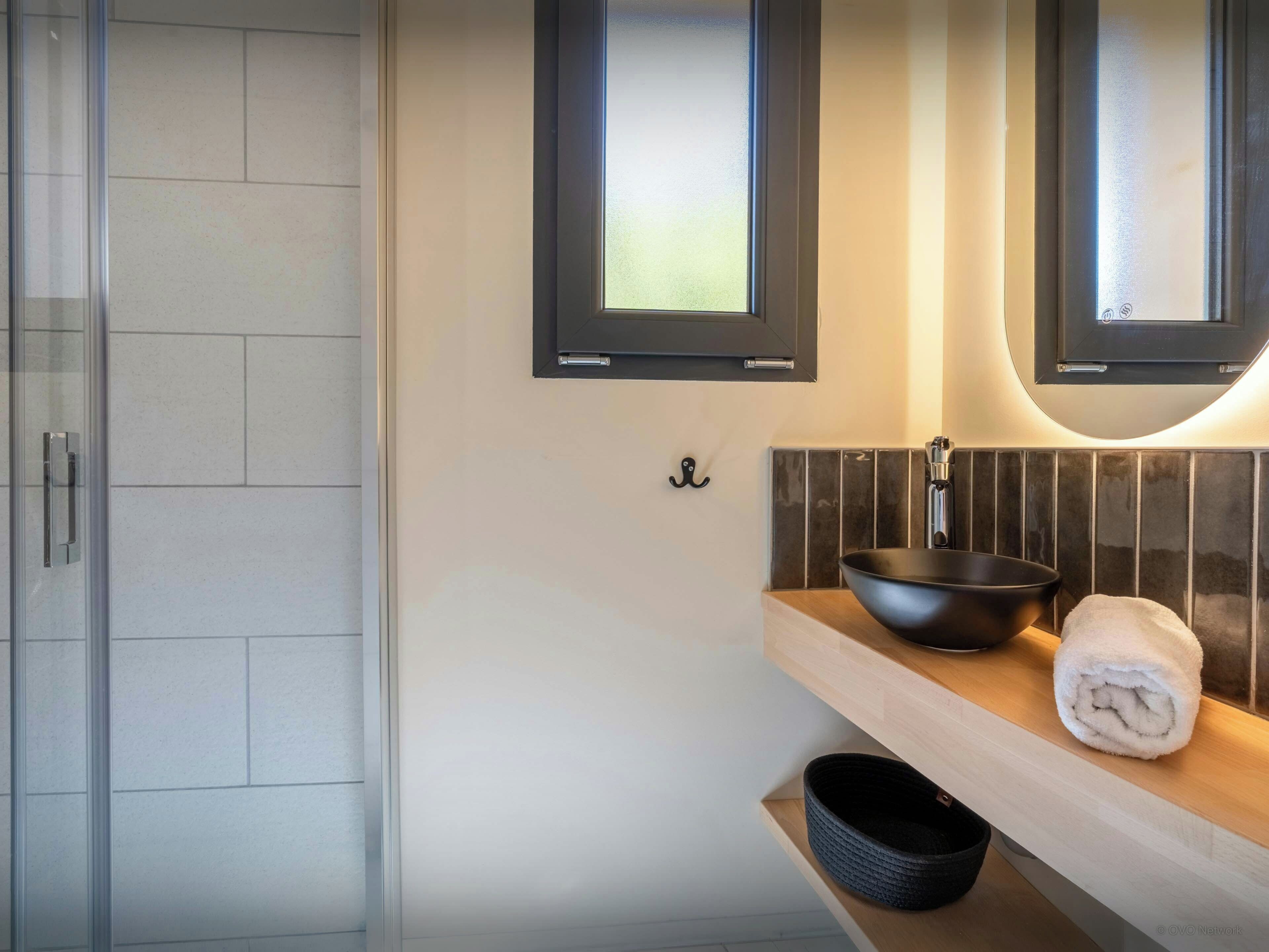 A modern bathroom with a glass shower enclosure, a black basin, and a towel on a wooden shelf.