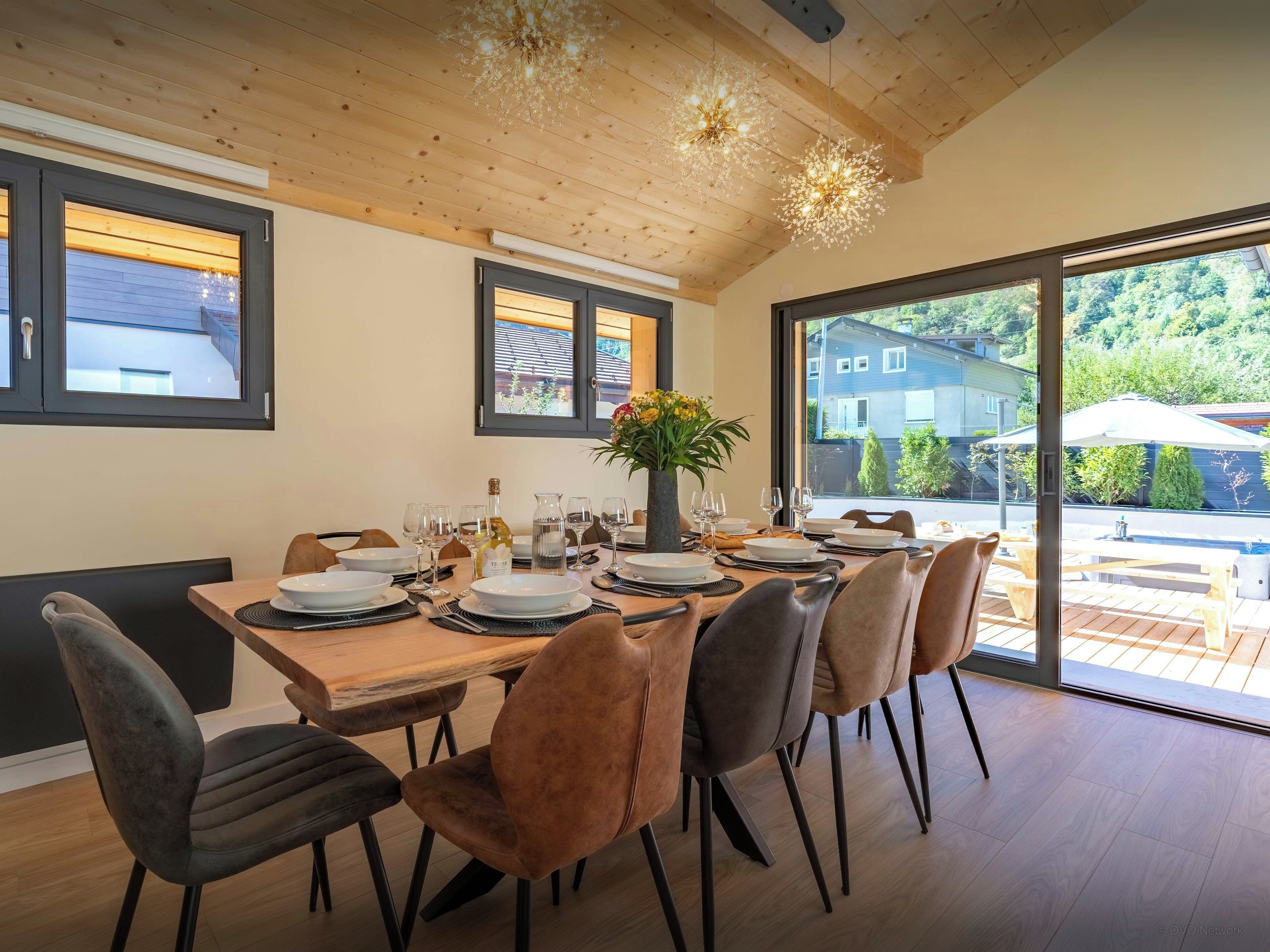 Un espace repas avec une table en bois dressée pour six personnes, entourée de chaises, et de grandes fenêtres donnant sur l’extérieur.