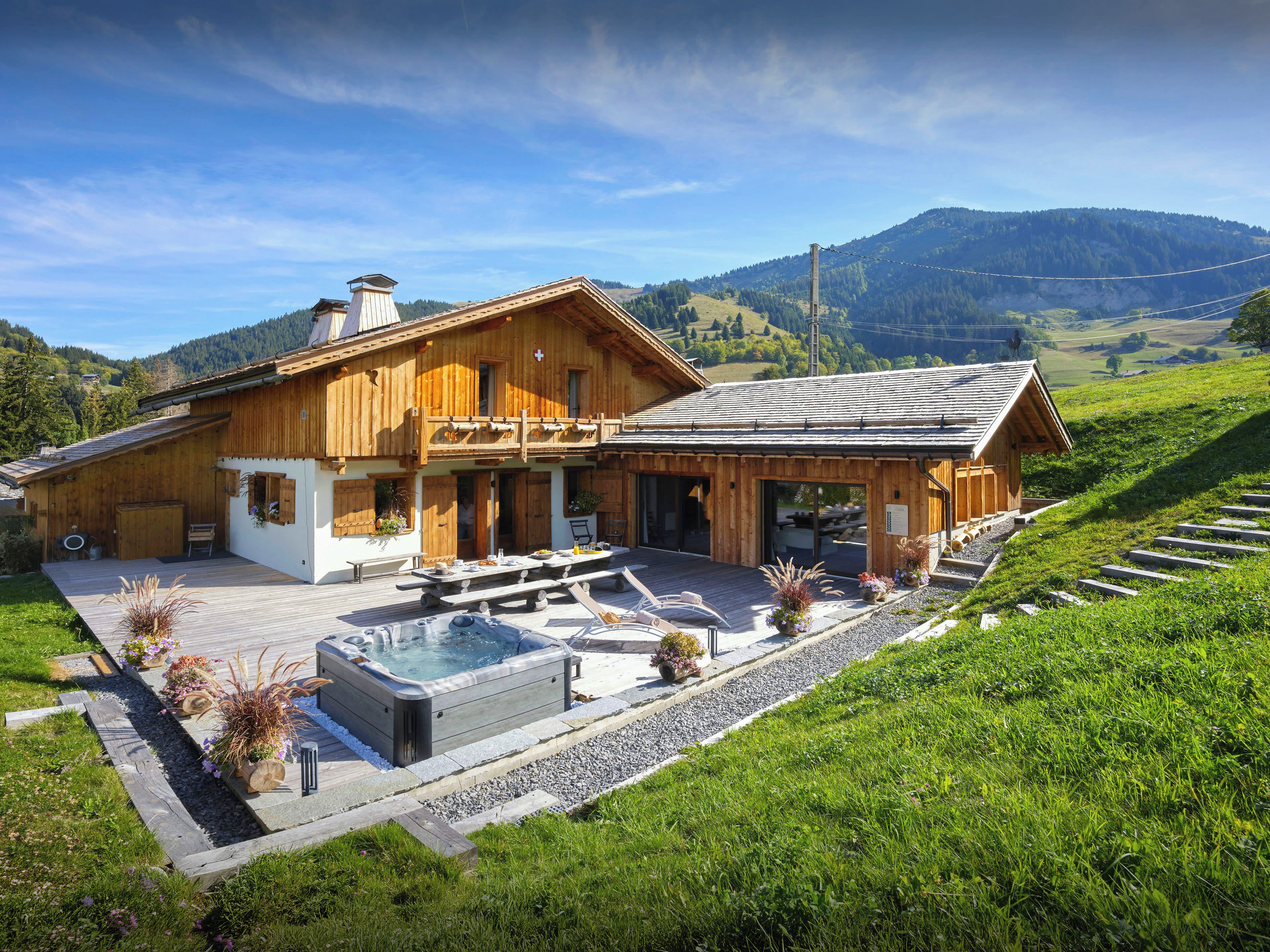 A wooden chalet with a hot tub, outdoor seating, and a view of the surrounding hills.
