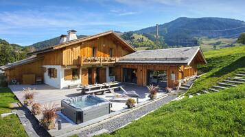 A wooden chalet with a hot tub, outdoor seating, and a view of the surrounding hills.