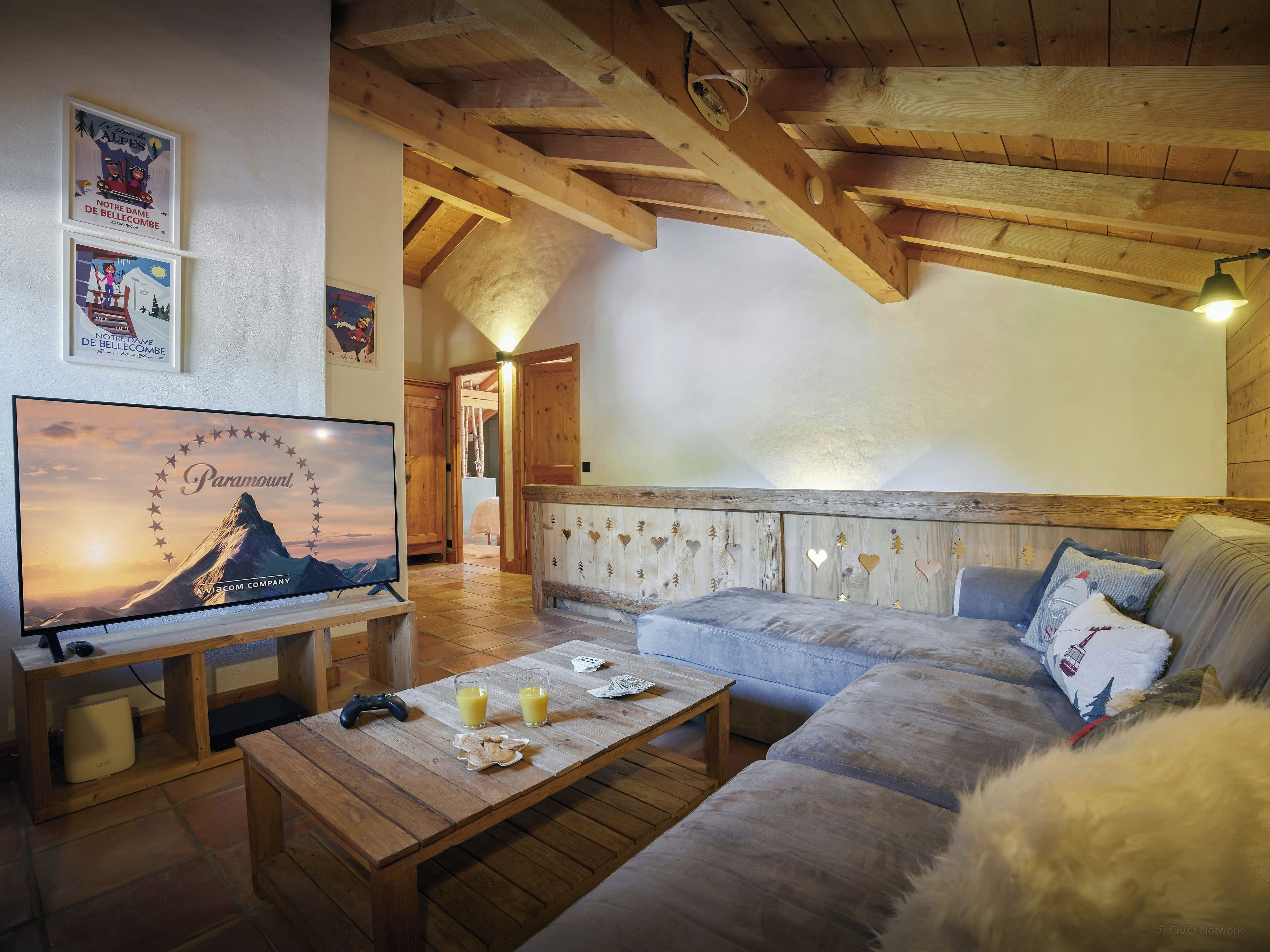 Ein gemütliches Wohnzimmer mit Holzbalkendecke, einem Flachbildfernseher auf einem Schrank, einer Couch und einem Couchtisch mit Getränken und einem Spielcontroller.