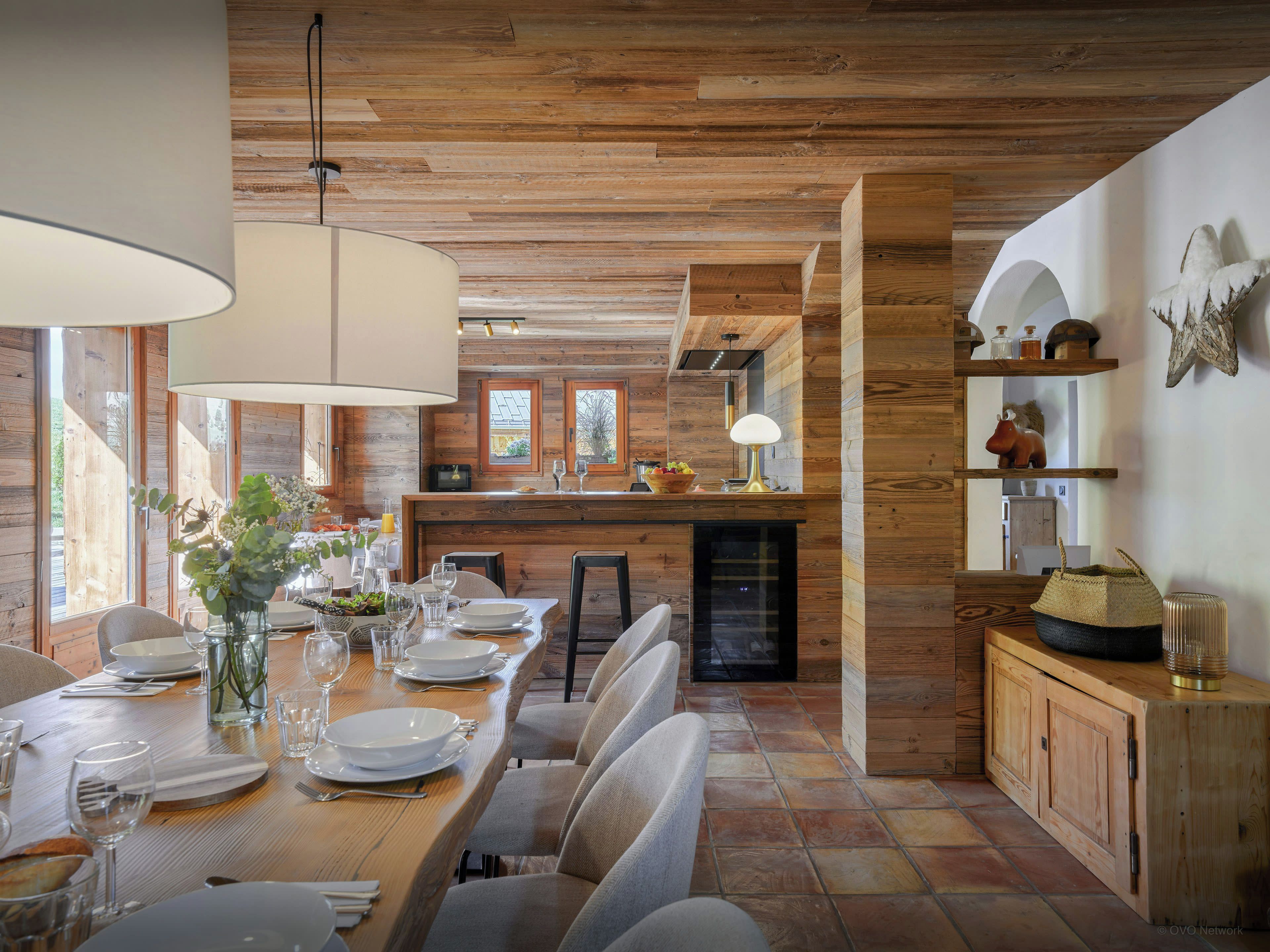 Ein Holzinterieur mit einem für vier Personen gedeckten Esstisch, einer Küchenzeile und einem großen Fenster, das natürliches Licht hereinlässt.