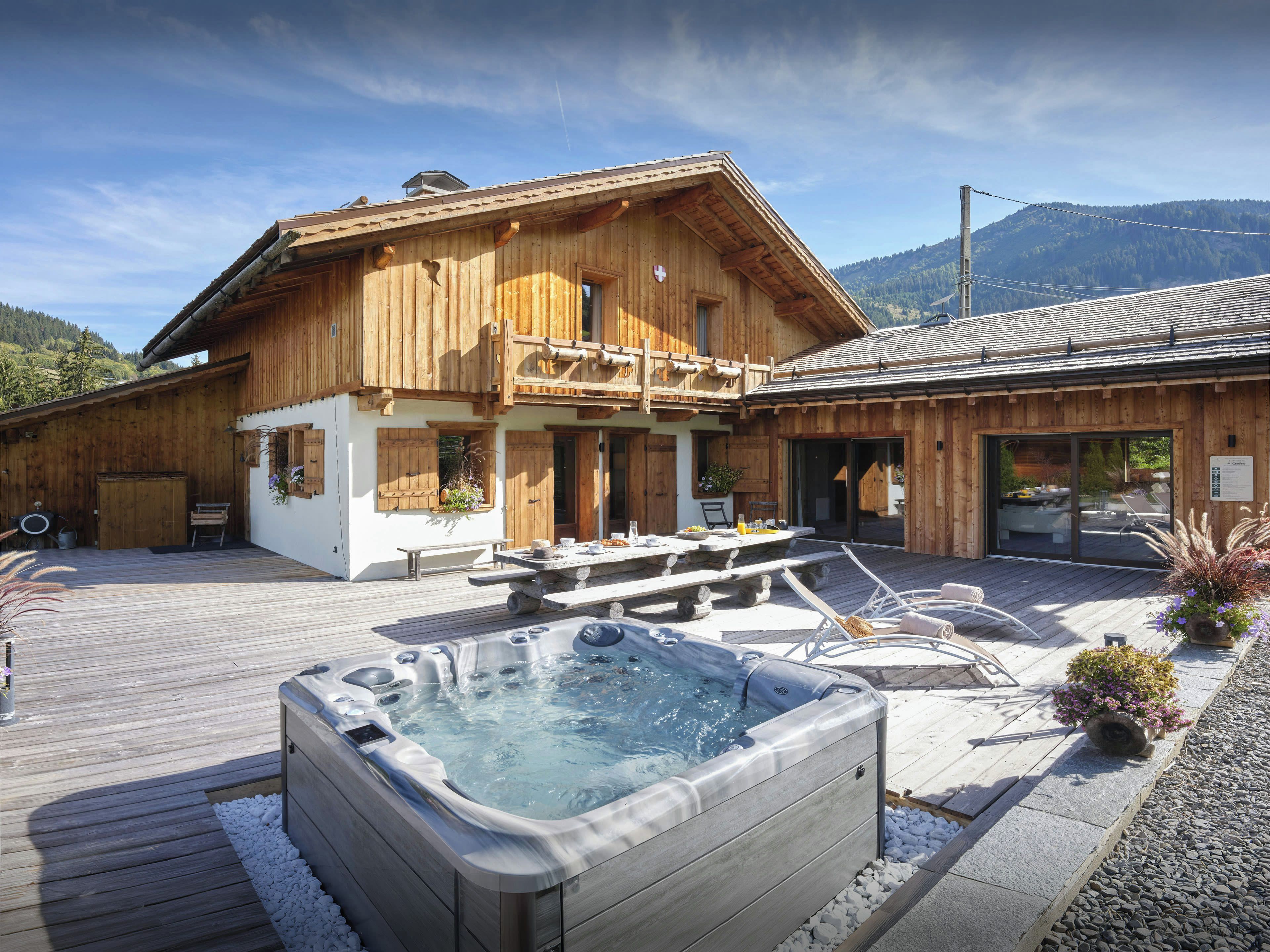 Un chalet in legno con idromassaggio, area salotto esterna e montagne sullo sfondo.