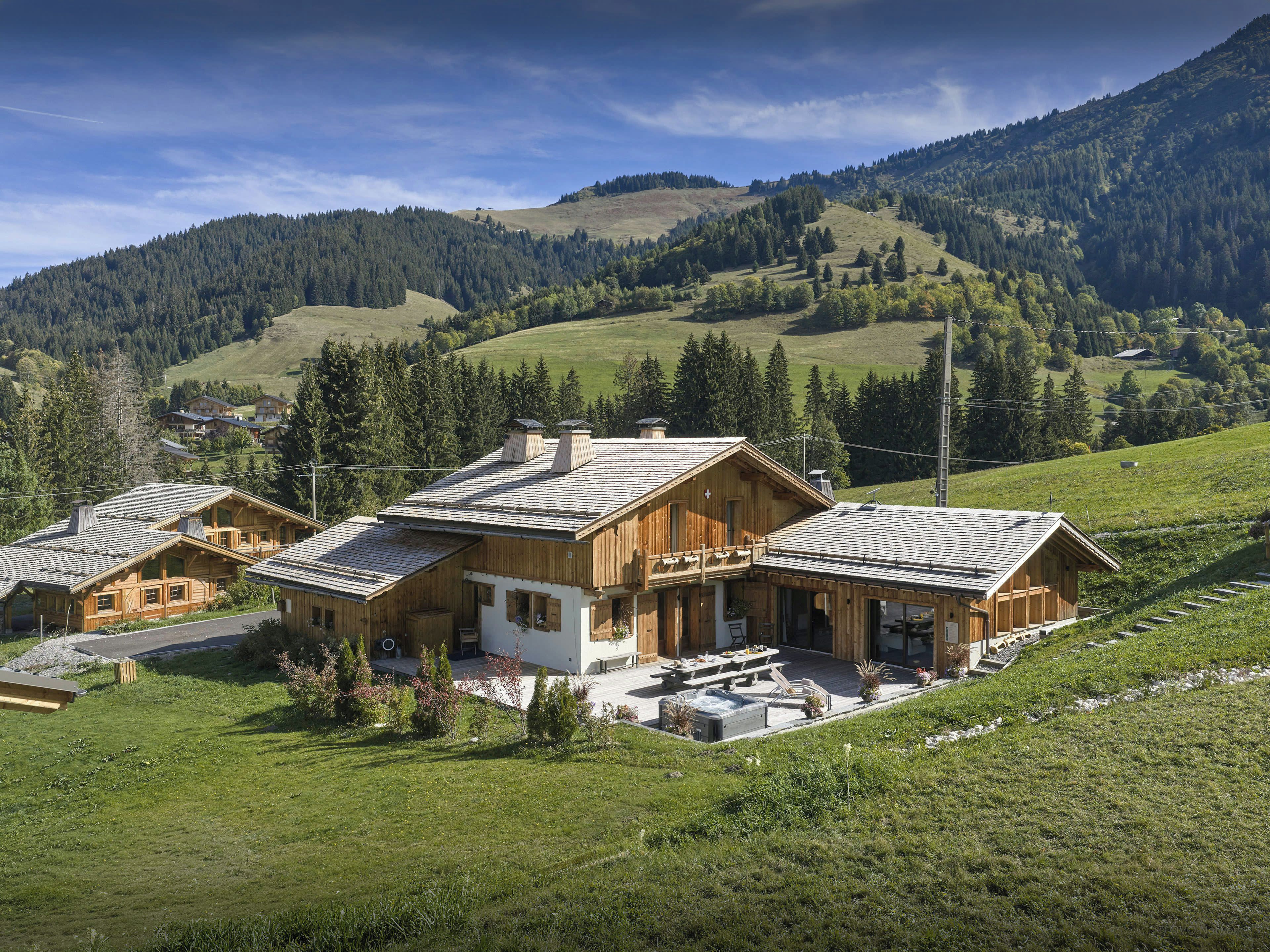 Eine Holzhütte mit Terrasse und Whirlpool, eingebettet in üppige, grüne Hügel unter blauem Himmel.