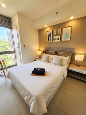 A hotel room with a large bed, two bedside tables with lamps, a headboard with artwork, and a view of greenery through the window. - Unixx south pattaya - 17th floor pool view modern all included condominium (ตำบล หนองปรือ)