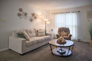 Een woonkamer met een beige bank, een rond bijzettafeltje met kaarsen, een vloerlamp en een stoel.