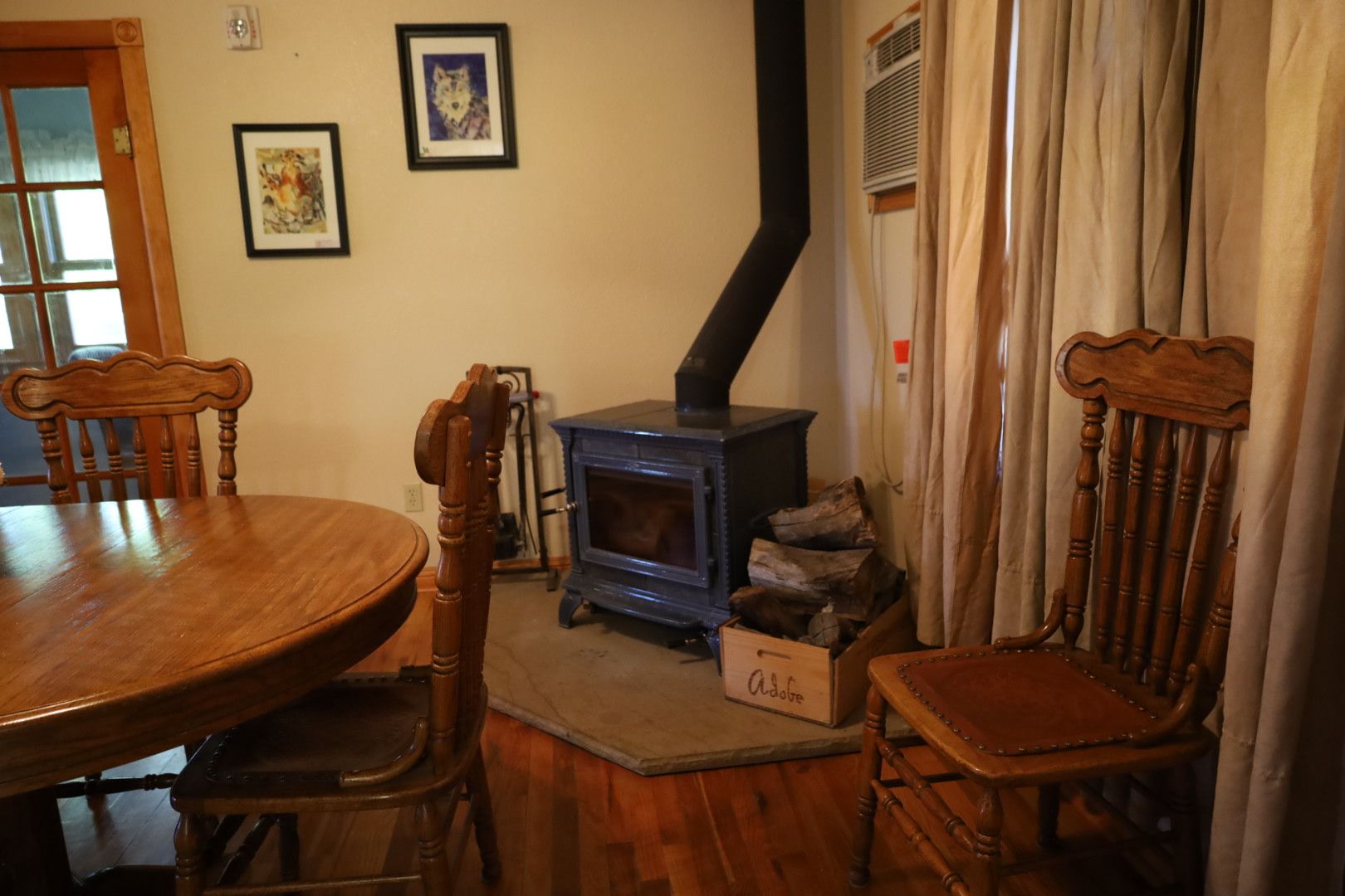 A dining area with a wooden table and chairs, a wood-burning stove, and a window with curtains.