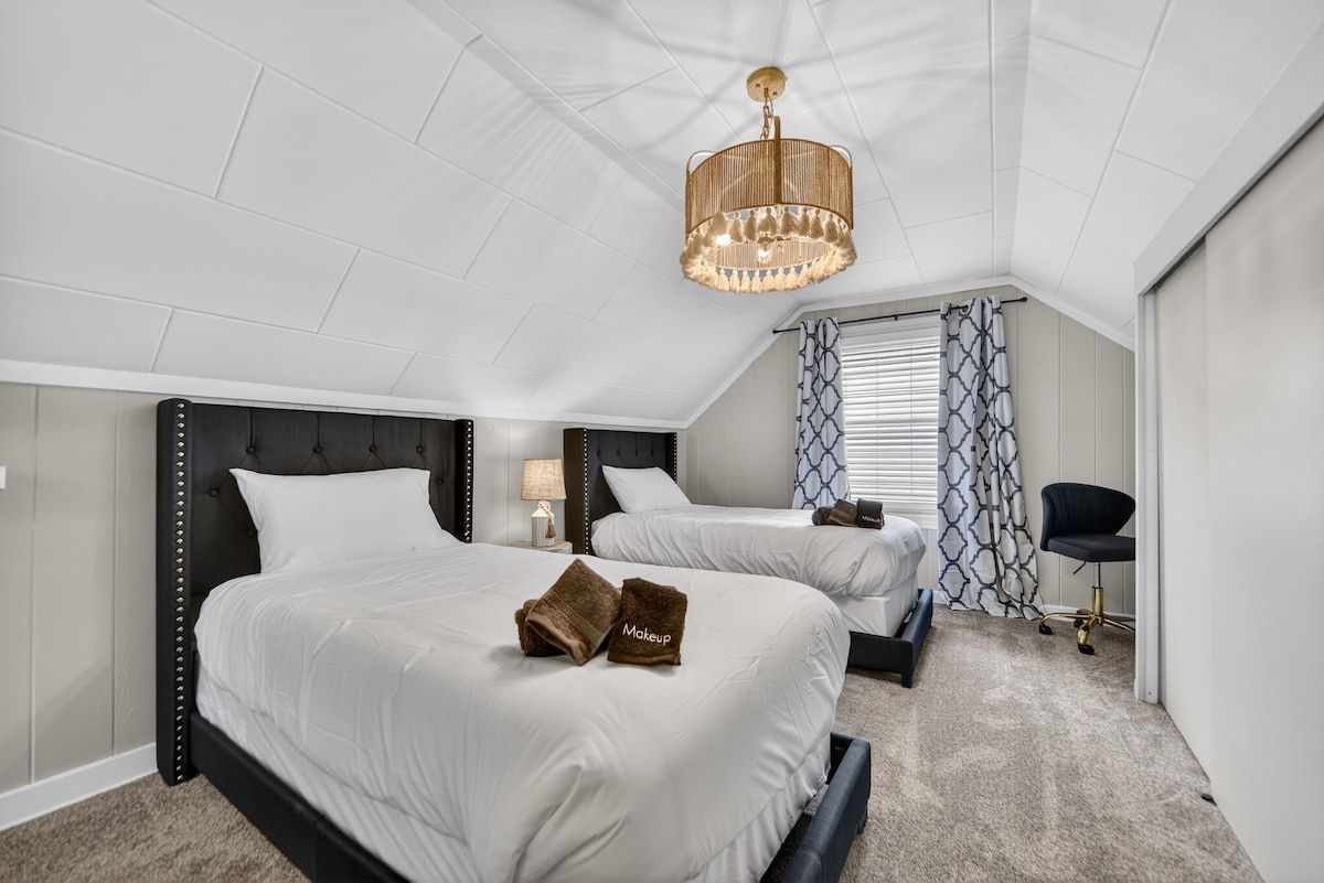 Une chambre d’hôtel avec deux lits, un lustre, une fenêtre avec des rideaux à motifs et une chaise.