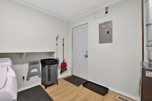 A clean, well-lit hallway with a white door, a utility meter, a trash bin, and a broom leaning against the wall. - Pleasant on Pheasant 2BR I 1 BA (Lansing)