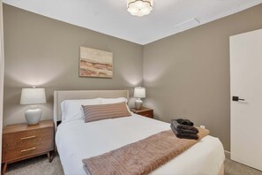A bedroom with a bed, two bedside tables with lamps, a nightstand, a door, and a painting on the wall. - Pleasant on Pheasant 2BR I 1 BA (Lansing)