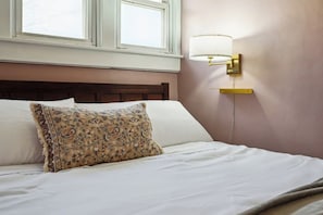 A bed with white linens and a floral-patterned pillow, a wall-mounted lamp, and a window above the bed. - Walk to Sparrow I 2 BR I 1 BA (Lansing)