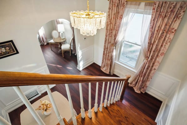 A staircase with a wooden handrail leading to a room with a chandelier, a window with floral curtains, and a white chair. - Walk to Sparrow I 2 BR I 1 BA (Lansing)