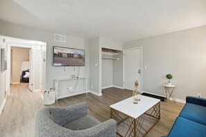 A modern living room with a grey armchair, a white coffee table, and a blue sofa. There is a flat-screen TV mounted on the wall, a small side table with a plant, and a white door leading to another room. - The Hangar #11 - 2BR - 1BA (Lansing)