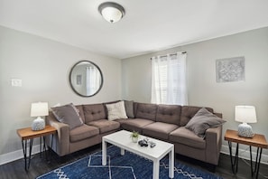 A modern living room with a sectional sofa, a round mirror, and two side tables with lamps. - Little Loft of Adventure | 1 Br 1 Ba (Lansing)