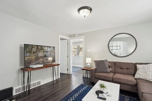 A living room with a flat-screen TV, a coffee table with a plant, a brown sofa, and a round mirror on the wall. - Little Loft of Adventure | 1 Br 1 Ba (Lansing)