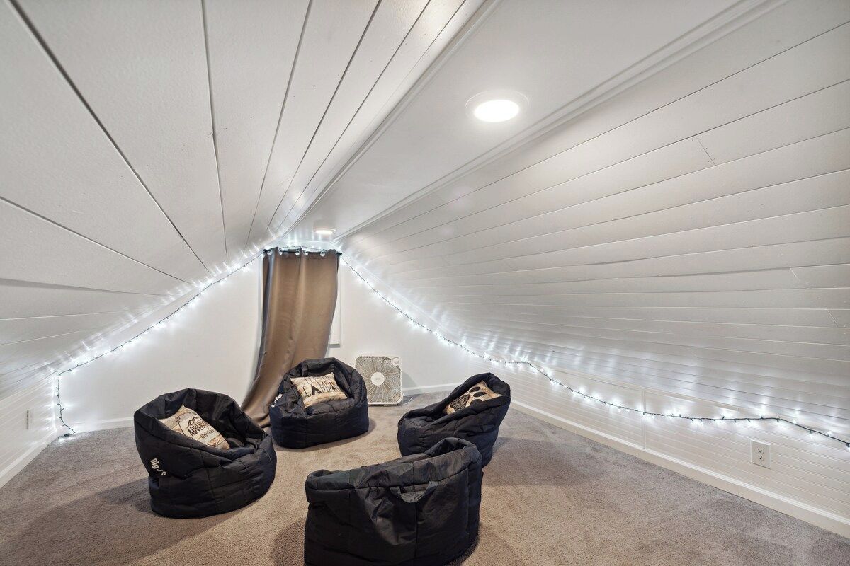 A narrow attic room with sloped ceilings, three bean bags, a fan, and string lights.