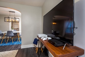 A modern living room with a large flat-screen TV, gaming console, and a wooden coffee table. - Little Loft of Adventure | 1 Br 1 Ba (Lansing)