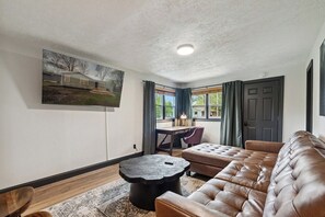 A living room with a leather sofa, a round coffee table, a desk with a chair, and a window with curtains. - 3BR/1BA w/Backyard & Firepit (Lansing)