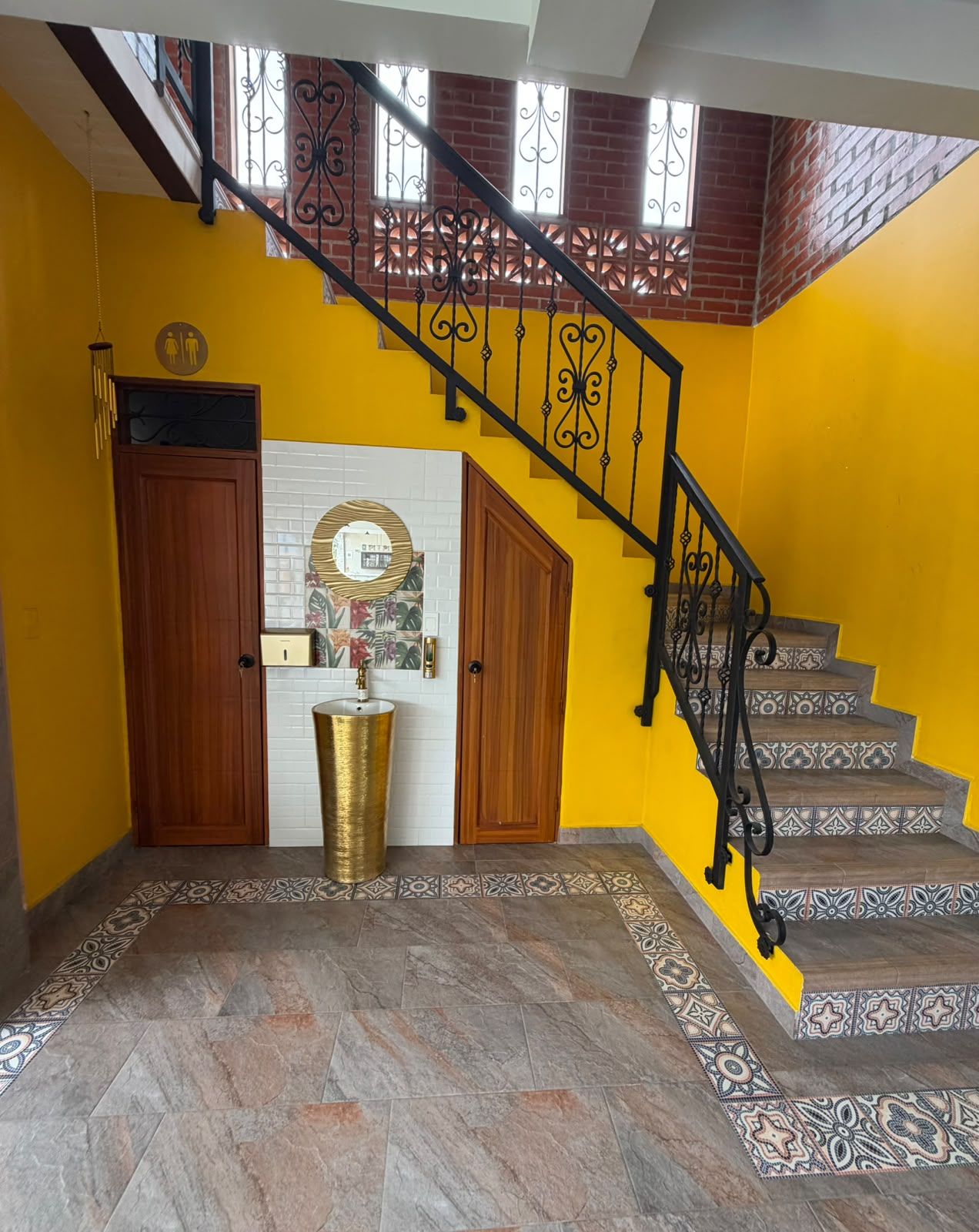 Una escalera con barandales negros ornamentales que conduce a un nivel superior, una puerta de madera, una pared de azulejos blancos con un espejo circular y baldosas de suelo con diseños.