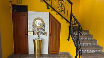 Una escalera con barandales negros ornamentales que conduce a un nivel superior, una puerta de madera, una pared de azulejos blancos con un espejo circular y baldosas de suelo con diseños.