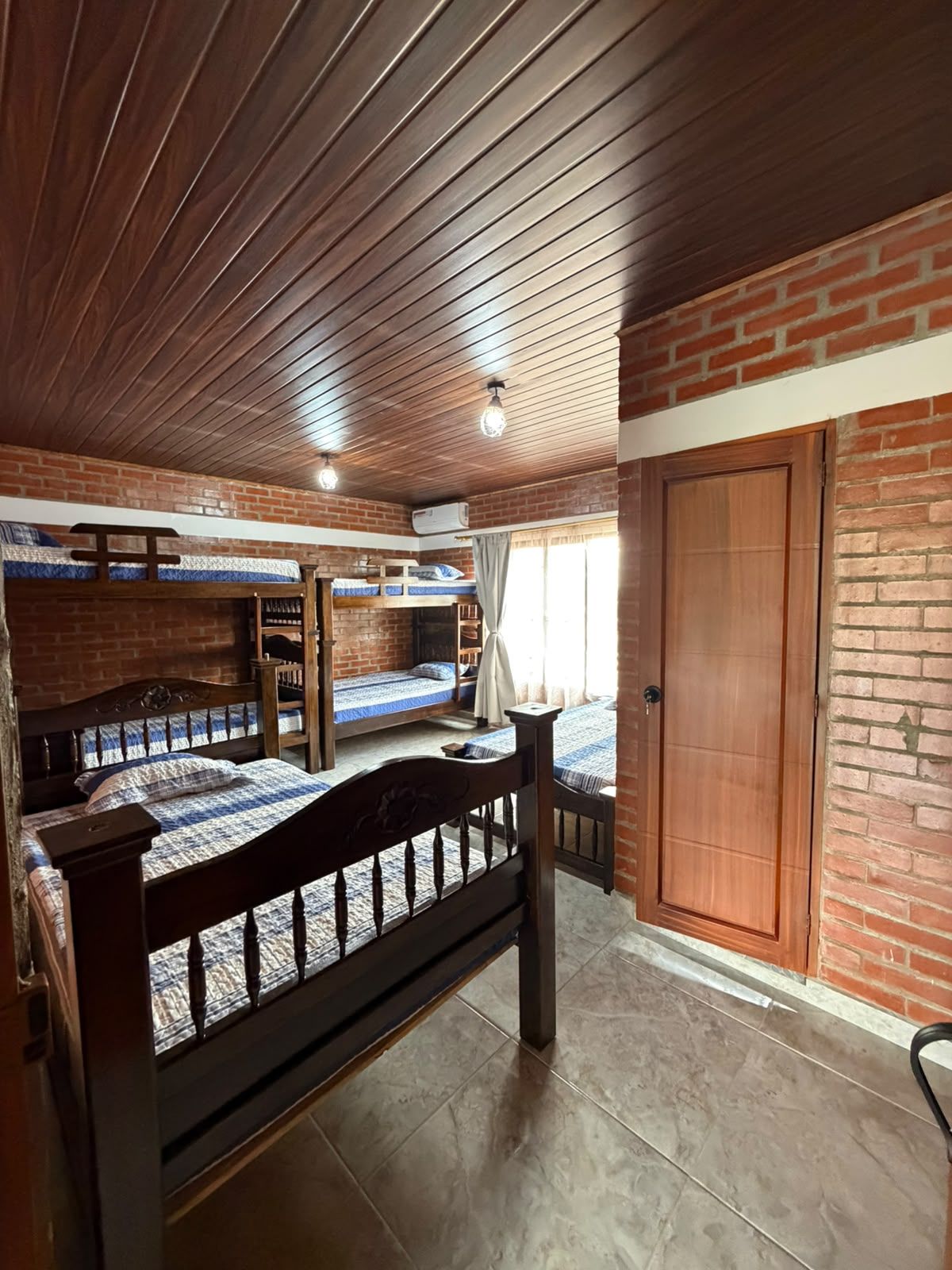 Una habitación con techo de madera, paredes de ladrillo, cama de madera, armario y ventana con cortinas.