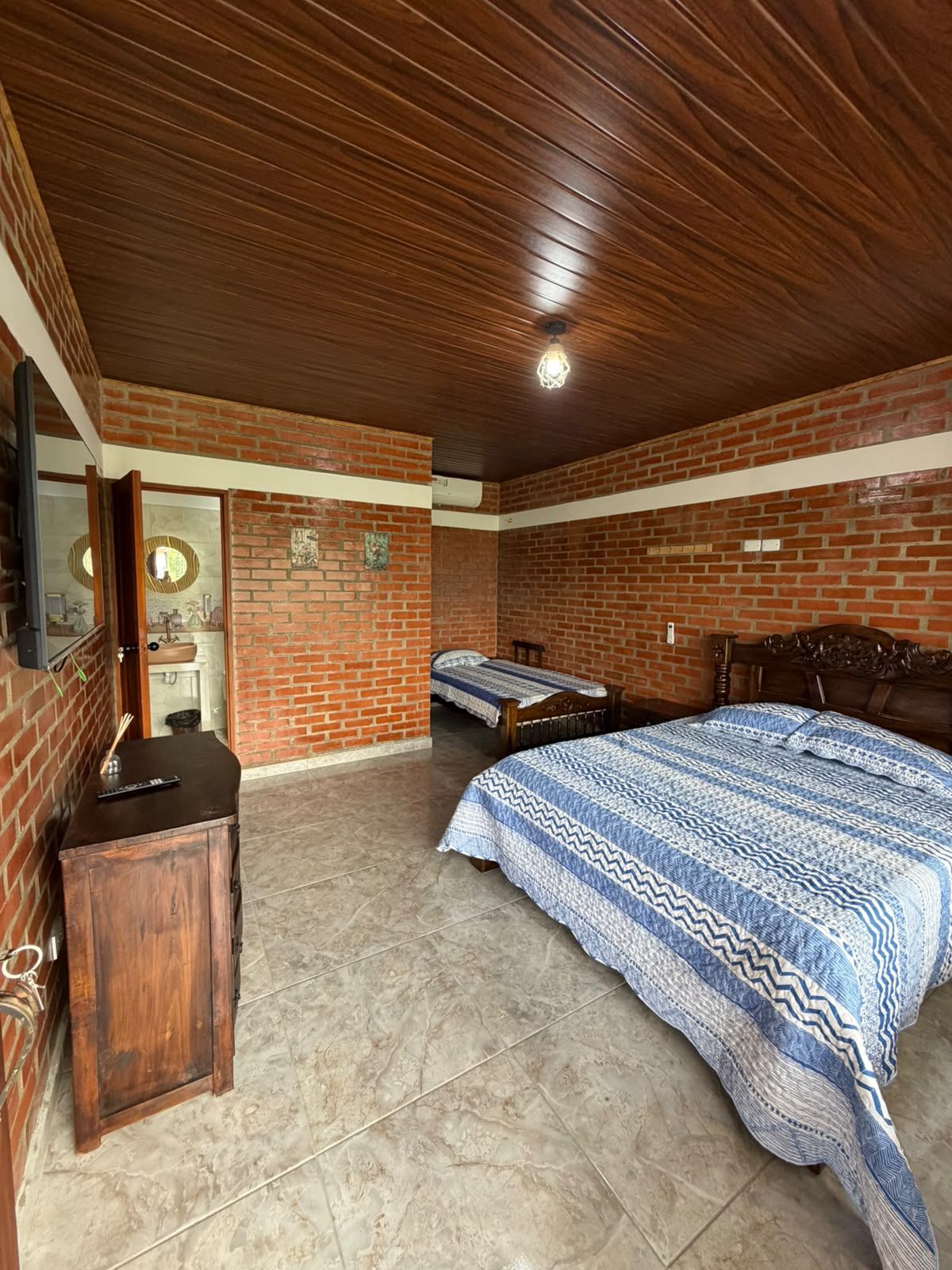 Une chambre avec un mur en briques, un plafond en bois, un lit recouvert d’une couvre-lit à motifs, une table de chevet et un miroir.